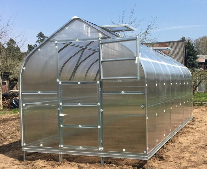 Clear polycarbonate greenhouse on a dirt field with trees and blue sky in the background