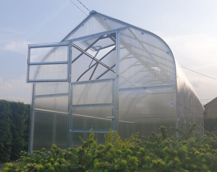 Greenhouse with transparent walls and a metal frame, surrounded by greenery.